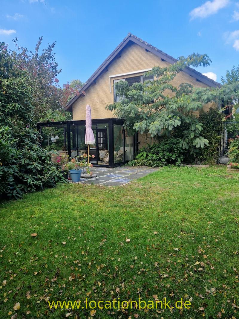 Haus mit steilem Satteldach und verglastem Anbau, umgeben von einem grünen Garten.