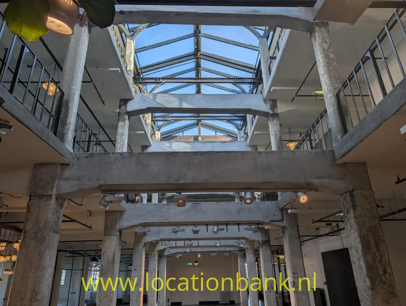 Betonnen draagbalken en zuilen kenmerken een interieur onder een glazen dak met metalen balustrades.