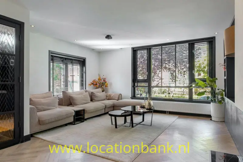 Woonkamer met een beige hoekbank, ronde salontafels en grote zwarte ramen met zicht op groen.