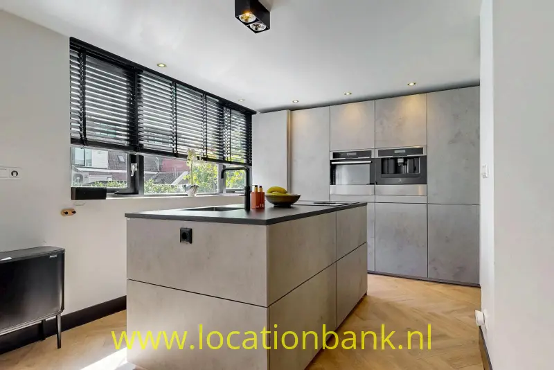 Keuken met een centraal kookeiland, hoge kasten, ingebouwde ovens en horizontale jaloezieën voor de ramen.