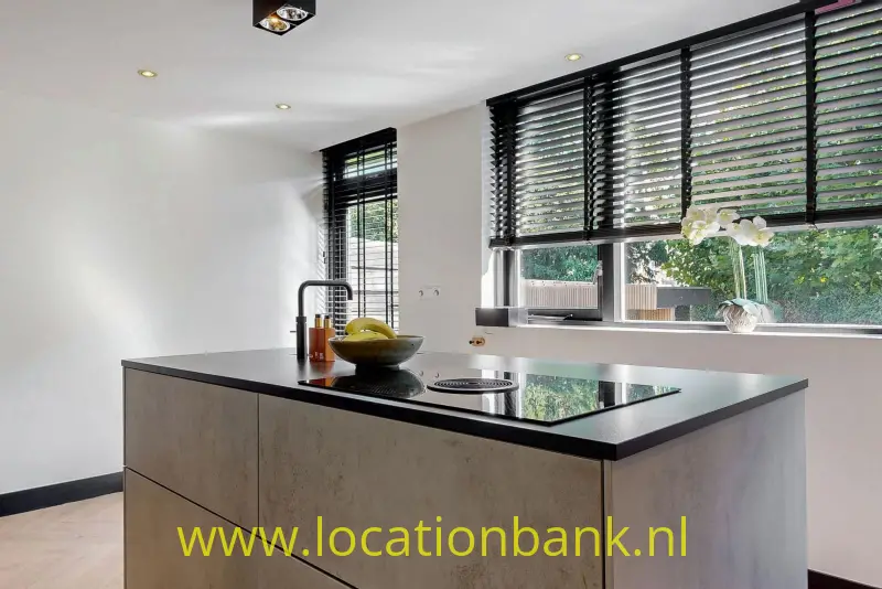 Keukeneiland met zwart werkblad, inductiekookplaat met ingebouwde afzuigiging, fruitschaal en grote ramen met jaloezieën.