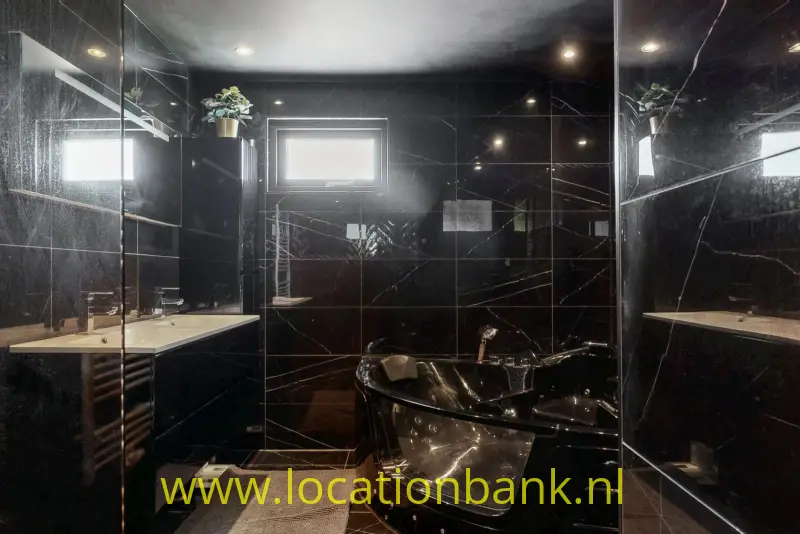 Een vrijstaand zwart bad staat in een badkamer met zwarte tegels, spiegels, een wastafel en een klein raam.