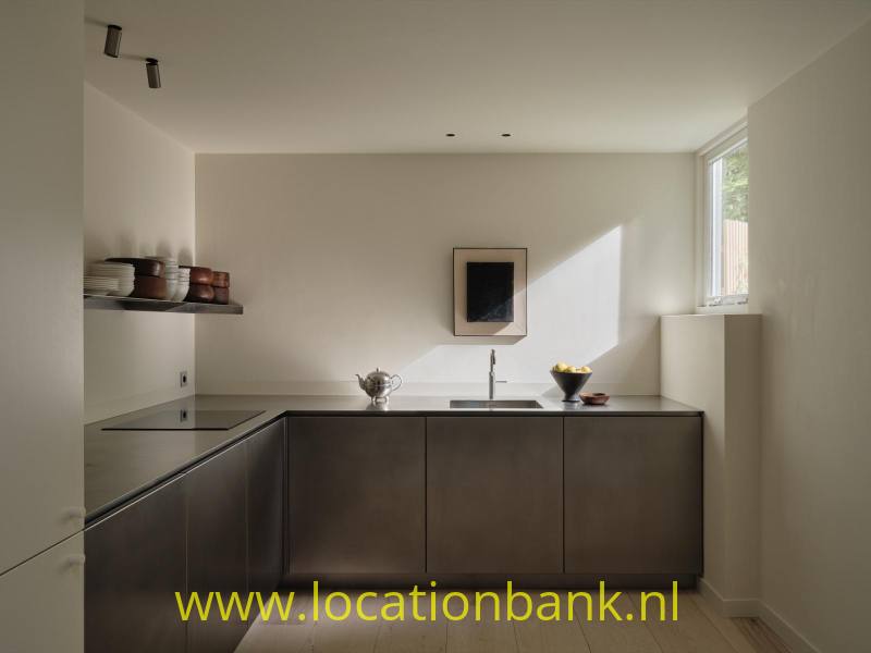 Minimalistische, moderne keuken met strakke donkere maatwerkfronten en een ingetogen, luxe uitstraling. Het natuurlijke licht valt mooi binnen via het zijraam en creëert een sfeervol contrast tussen licht en donkere materialen. Geschikt voor lifestyle-, food-, interieur- en commercials.