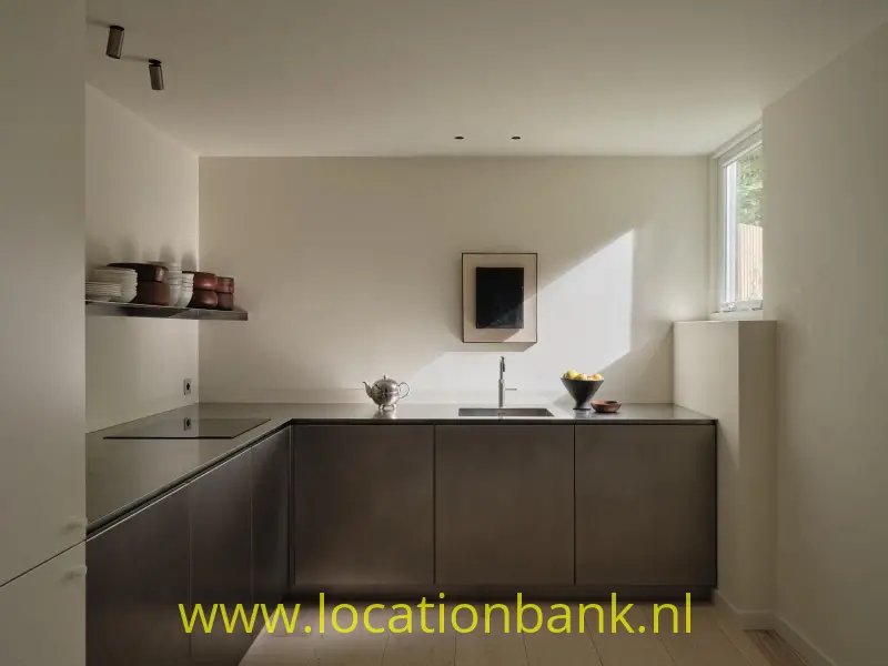 Minimalistische, moderne keuken met strakke donkere maatwerkfronten en een ingetogen, luxe uitstraling. Het natuurlijke licht valt mooi binnen via het zijraam en creëert een sfeervol contrast tussen licht en donkere materialen. Geschikt voor lifestyle-, food-, interieur- en commercials.