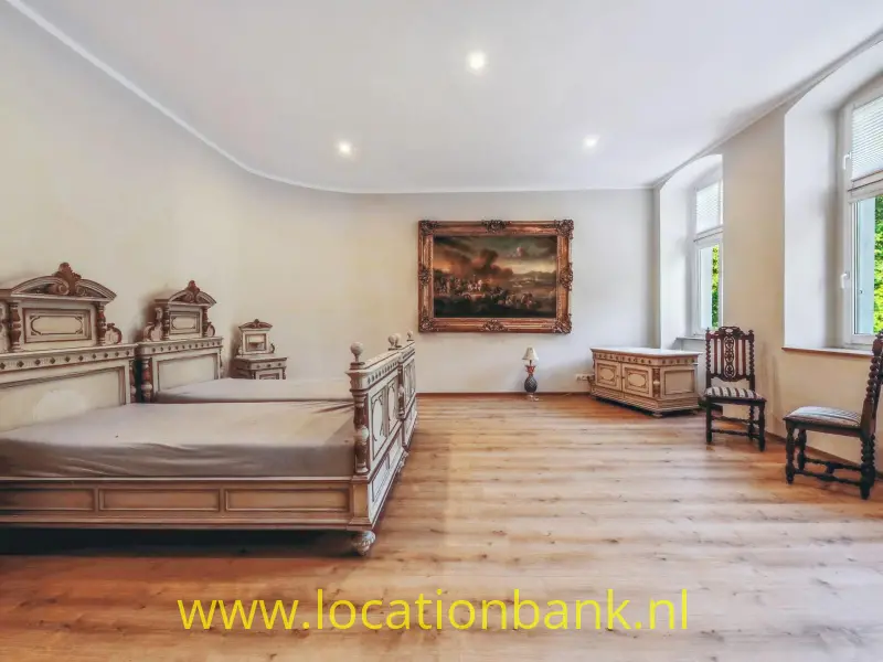 Indrukwekkende, zeer ruime slaapkamer met een uitgesproken klassiek landhuis-karakter. Het grote, rijk gedecoreerde houten bed vormt het visuele middelpunt van de ruimte. De antieke of antiek aandoende meubels, klassieke schilderijen en hoge ramen versterken de sfeer van een statig Europees interieur.

De combinatie van de royale maatvoering, hoge plafonds, lichte wanden en warme houten vloer zorgt voor een rustige maar tegelijkertijd zeer cinematografische ruimte. Het interieur heeft een tijdloze uitstraling en kan zowel historisch als hedendaags worden ingezet.

Bijzonder geschikt voor films, series, kostuumdrama, historische producties, luxury lifestyle, fashion- en interieurfotografie. De ruimte biedt veel mogelijkheden voor wide shots waarin de architectuur en het meubilair samen het beeld bepalen, maar ook voor meer intieme close-ups en portretten. De grote ramen zorgen voor een natuurlijke lichtbron en kunnen een zachte, schilderachtige sfeer creëren.