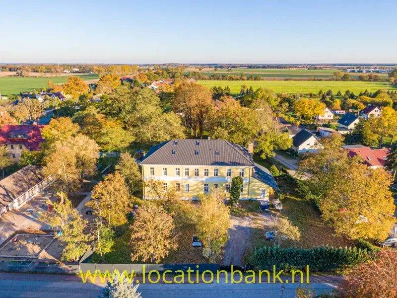 Een groot geel landhaus omringd door bomen en groene velden.