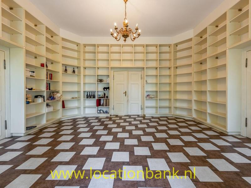 Karakteristieke bibliotheekkamer met kamerhoge witte boekenkasten, klassieke dubbele deuren en een opvallende vintage kroonluchter. Het bijzondere tegelpatroon en de symmetrische inrichting geven deze ruimte een authentieke, statige uitstraling. Ideaal als filmlocatie voor historische scènes, interieurfotografie en stijlvolle producties.