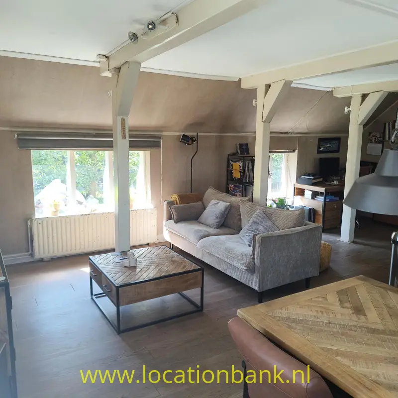 Woonkamer met een grijze hoekbank, houten salontafel, zichtbare draagbalken en ramen met uitzicht op groen.