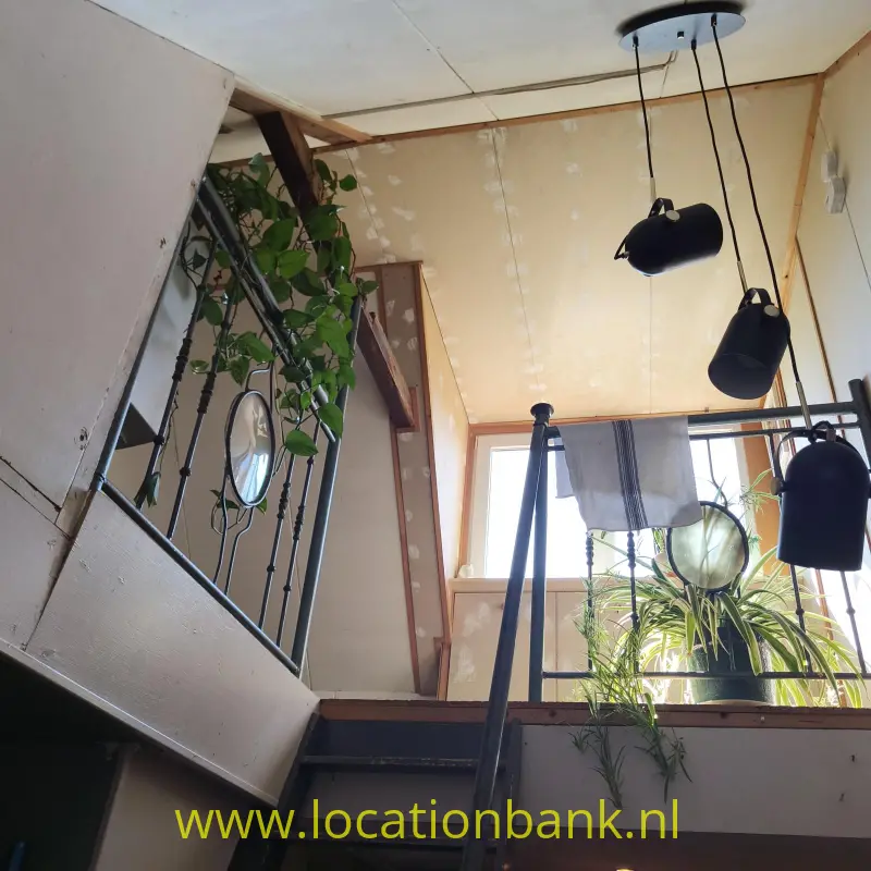Een trap met een zwarte metalen leuning, hanglampen, kamerplanten en grote ramen in een lichte binnenruimte.