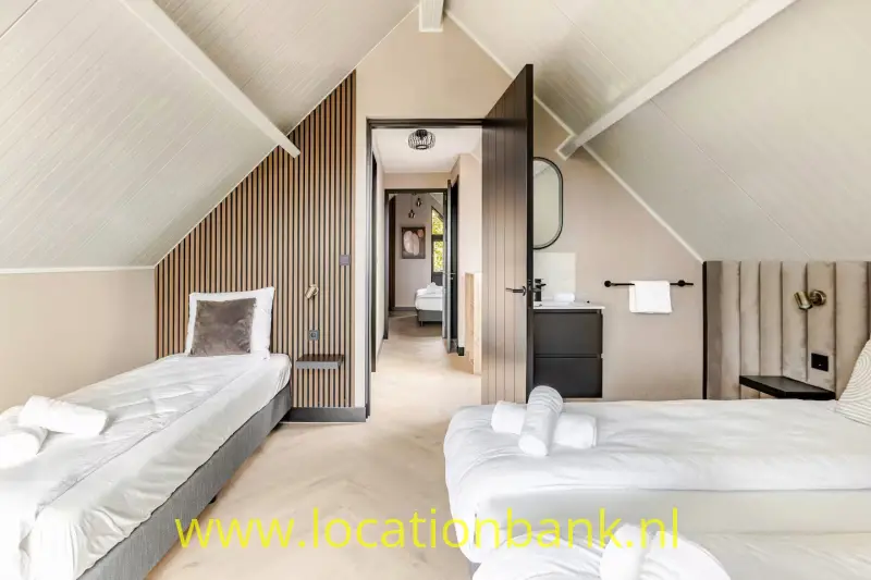 Twee opgemaakte witte bedden staan in een slaapkamer met een schuin plafond en een openstaande deur.