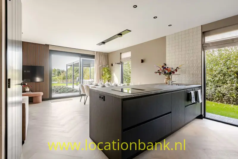 Groot donker keukeneiland met kookplaat staat centraal in een lichte keuken met glazen deuren naar de tuin.