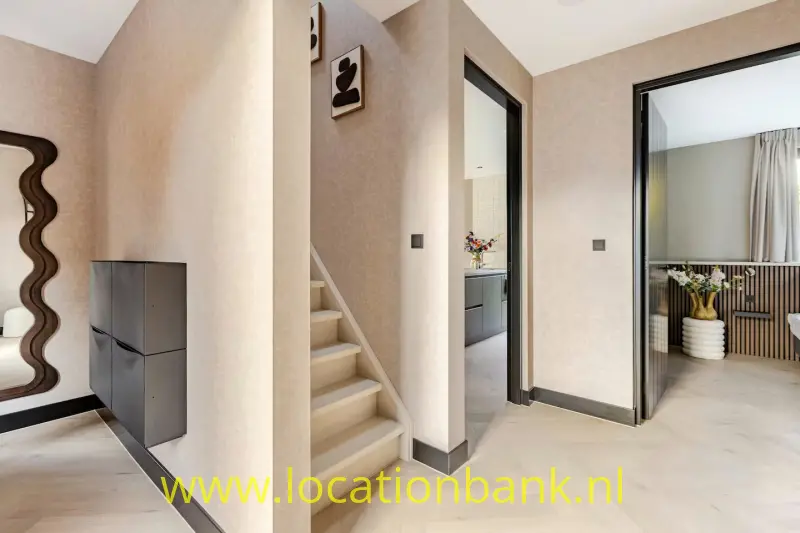 Een lichte hal met trap naar boven, een donker wand meubel en twee open deuren.