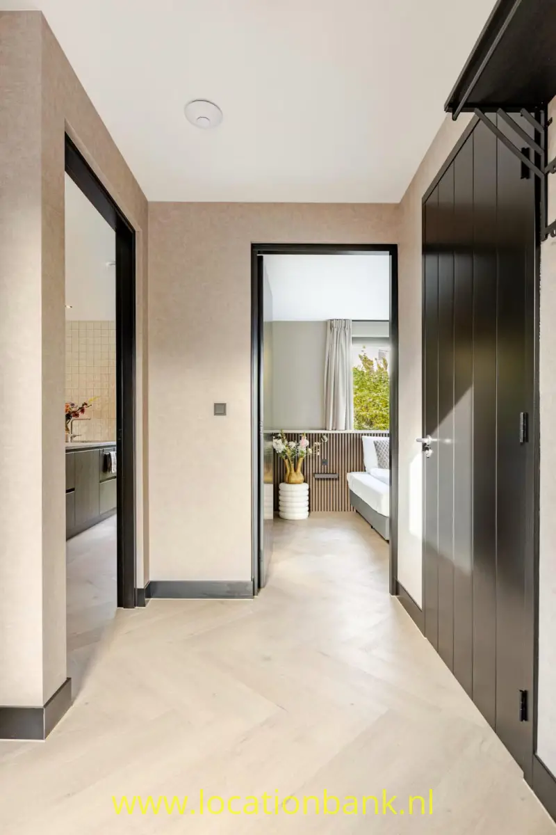 Lichte hal met een visgraatvloer, donkere deuren en een open doorgang naar een slaapkamer