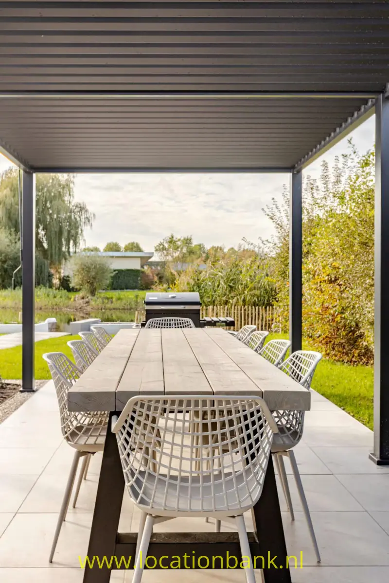Overdekt tuin terras met lange houten eettafel en gevlochten stoelen, omgeven door een groene tuin.
