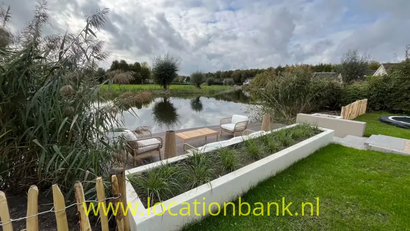 Gazon en verhoogde witte plantenbedden aan een vijver, met een houten terras, stoelen en hoog riet.