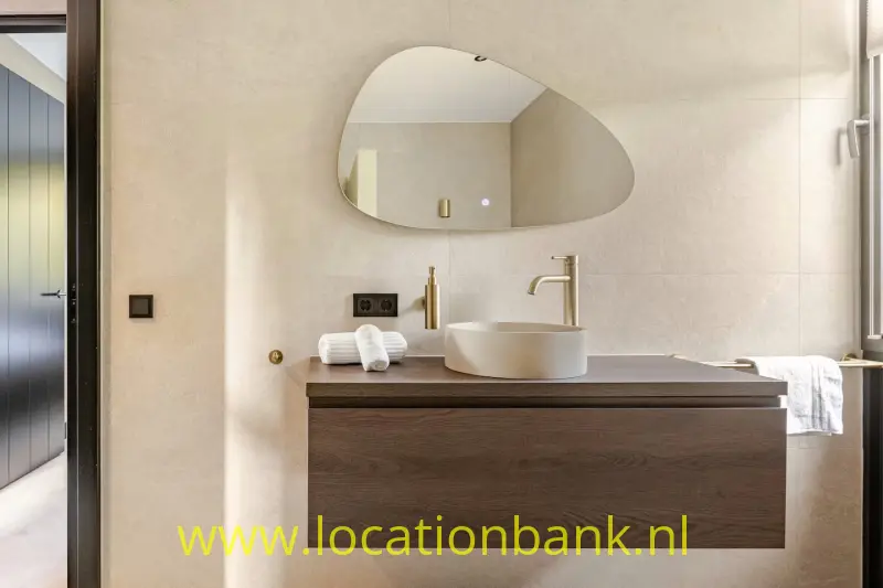 Wastafelmeubel met een ronde waskom, asymmetrische spiegel en witte handdoek tegen een betegelde wand.