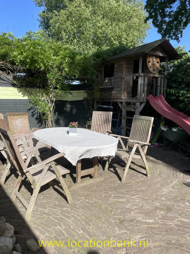Een houten speelhuisje met rode glijbaan staat achter een tuintafel met wit tafelkleed op een geplaveid terras.