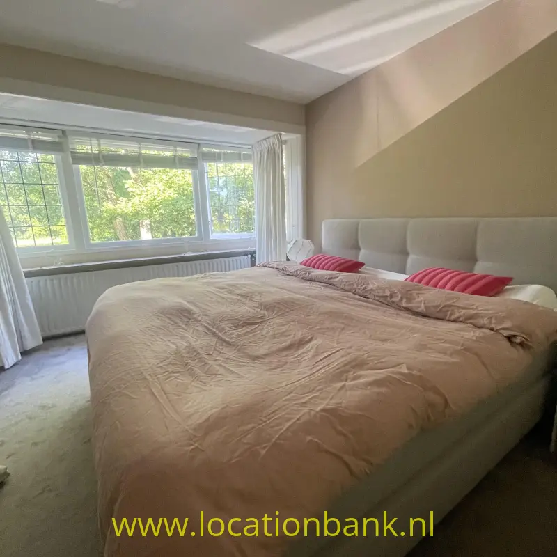 Een groot bed met een beige dekbed, roze kussens en een gestoffeerd hoofdbord staat voor een groot raam.