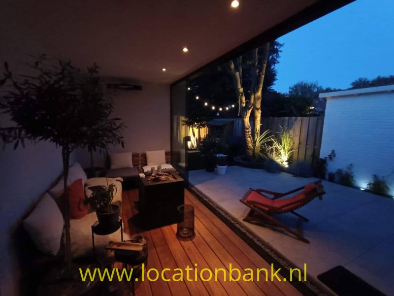 Overdekte patio met zithoek, houten vlonder, planten en rode ligstoelen naast een betegelde binnenplaats.