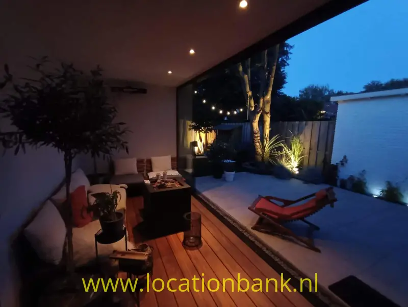 Overdekte patio met zithoek, houten vlonder, planten en rode ligstoelen naast een betegelde binnenplaats.