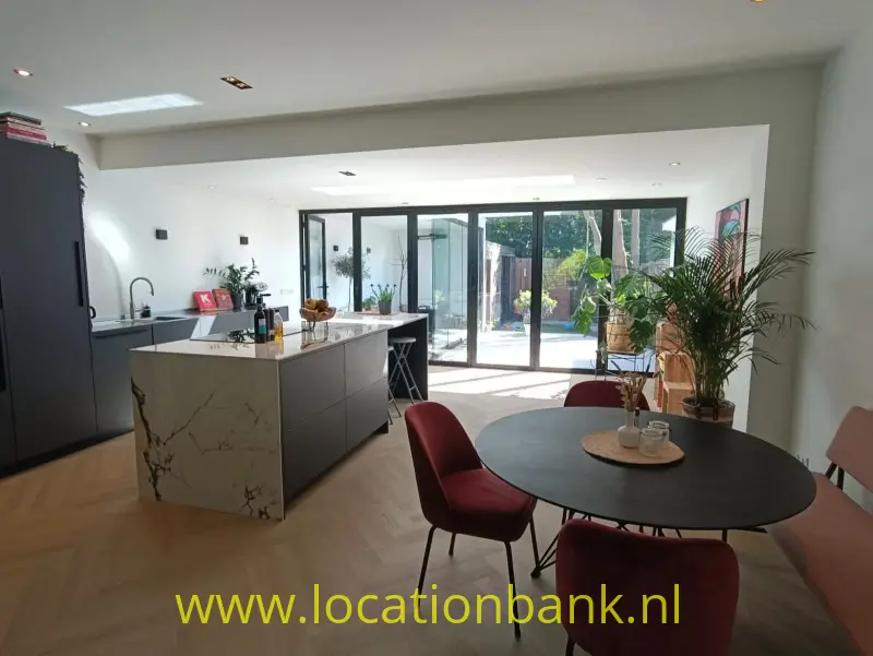 Open keuken met kookeiland en ronde eettafel bij glazen deuren naar een tuin.