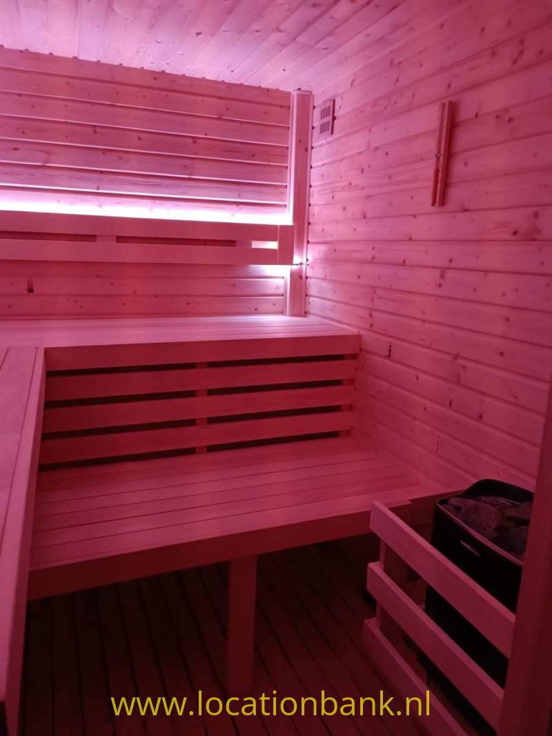 Houten sauna met meerdere zitbanken, horizontale wandplanken en een klein raam onder roze verlichting.