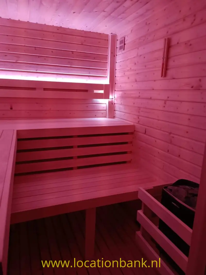 Houten sauna met meerdere zitbanken, horizontale wandplanken en een klein raam onder roze verlichting.
