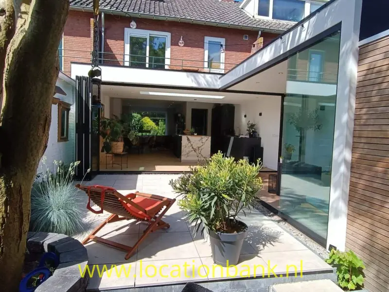 Binnenplaats met een betegeld terras, rode ligstoelen, plantenbakken en een glazen serre naast een bakstenen gevel.