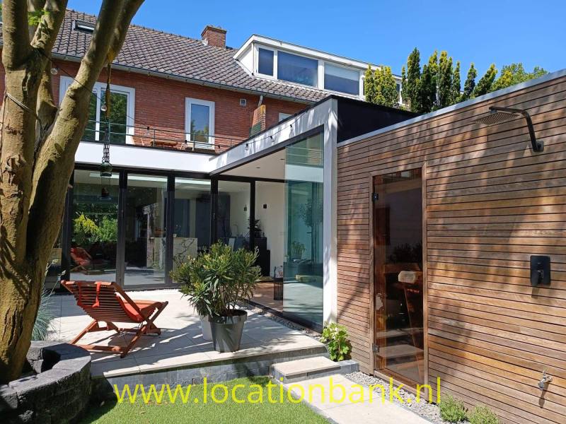 Bakstenen woning met glazen aanbouw, houten bijgebouw en patio met planten en twee ligstoelen.