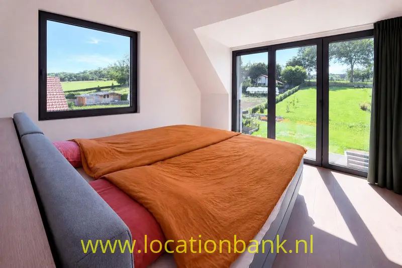 Tweepersoonsbed met oranje beddengoed naast grote ramen met uitzicht op een groen tuin landschap.