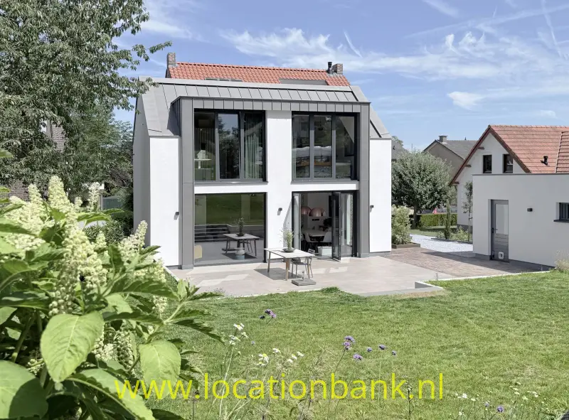 Moderne woning met grote glaspartijen, terras en aangelegde tuin naast een wit huis met rode dakpannen.