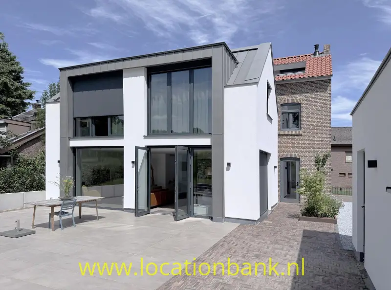 Witte woning met donkere gevelpanelen, grote ramen, een terras en een aangrenzend bakstenen gebouw.