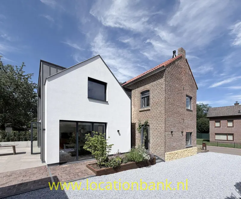 Witte moderne aanbouw naast een bakstenen huis, met grindterrein en plantenbakken langs de gevel.