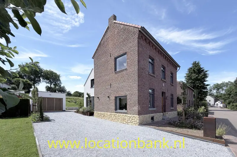 Een bakstenen huis met twee verdiepingen staat aan een brede grindoprit, naast andere woningen.