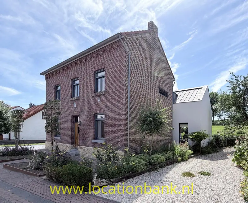 Bakstenen huis met een witte aanbouw, een grindpad en een aangelegde voortuin.