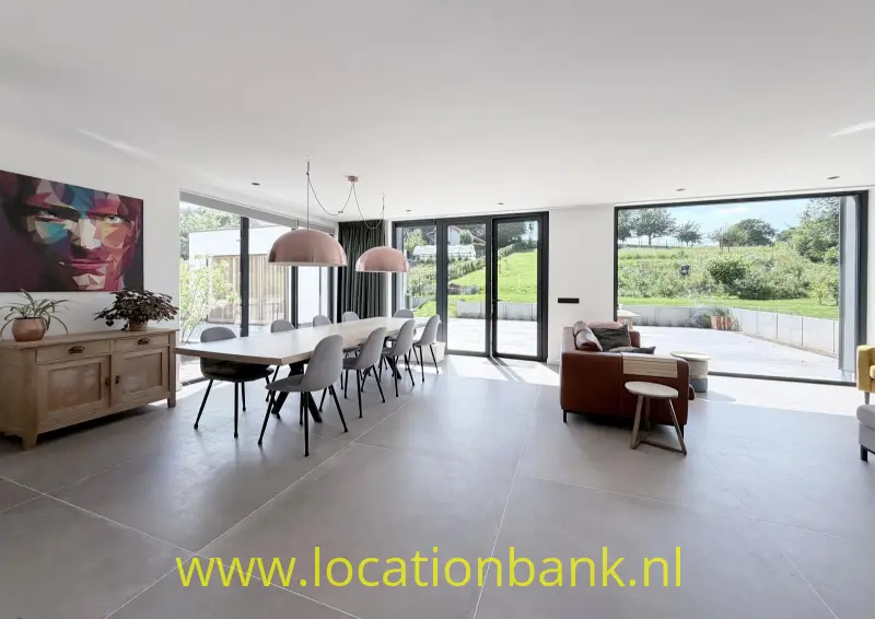 Open woon- en eetgedeelte met een lange eettafel, hanglampen en grote ramen op het groen.