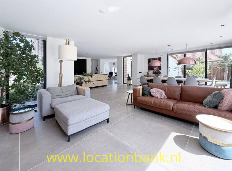 Ruime woonkamer met een grijze fauteuil, bruine bank, grote ramen en een open keuken.