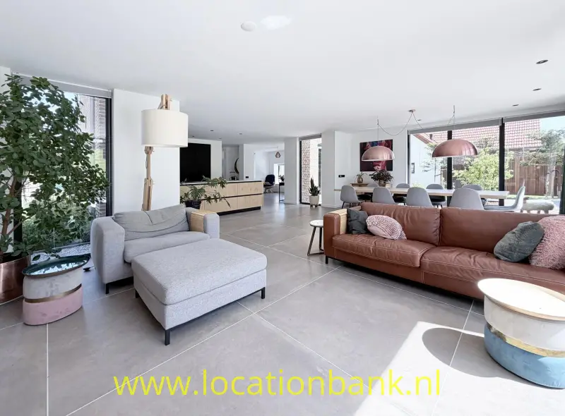 Ruime woonkamer met een grijze fauteuil, bruine bank, grote ramen en een open keuken.