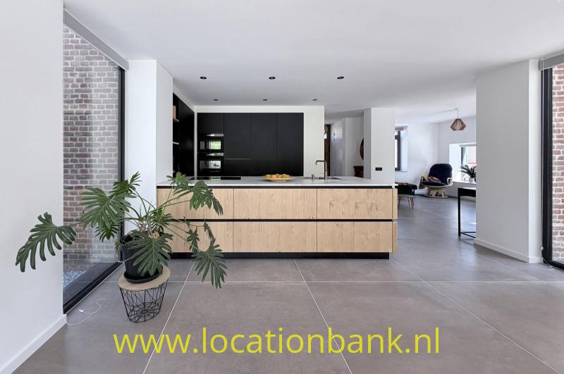 Open keuken met een groot houten kookeiland, zwarte kasten, een kamerplant en een bakstenen muur.