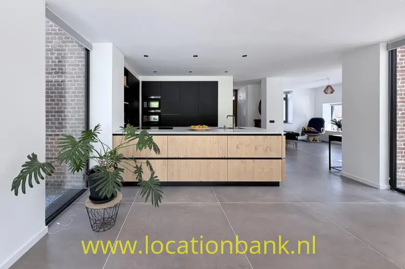 Open keuken met een groot houten kookeiland, zwarte kasten, een kamerplant en een bakstenen muur.