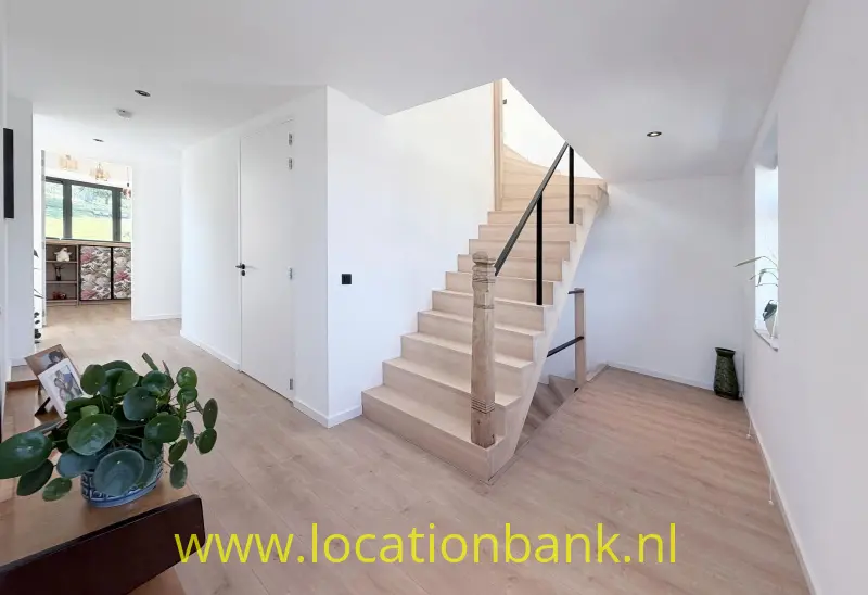 Houten trap met zwarte leuning in een lichte hal met houten vloer en kamerplanten.