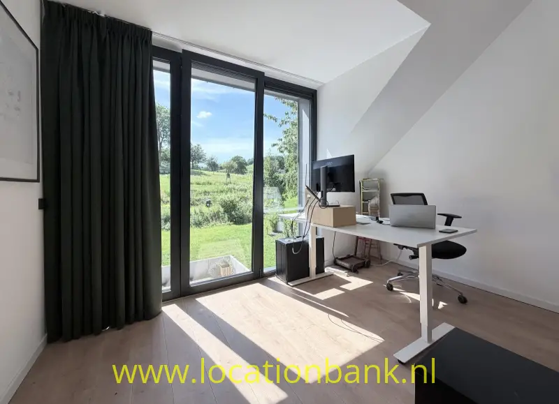 Werkplek onder een schuin plafond met bureau, monitor, laptop en hoge ramen met uitzicht op groen.