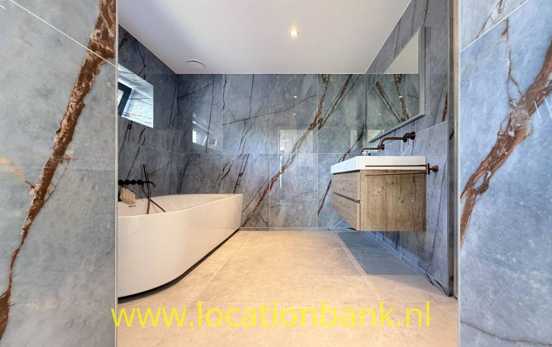 Badkamer met een vrijstaand wit bad, een houten wastafelmeubel en grijze wandtegels met bruine aders.