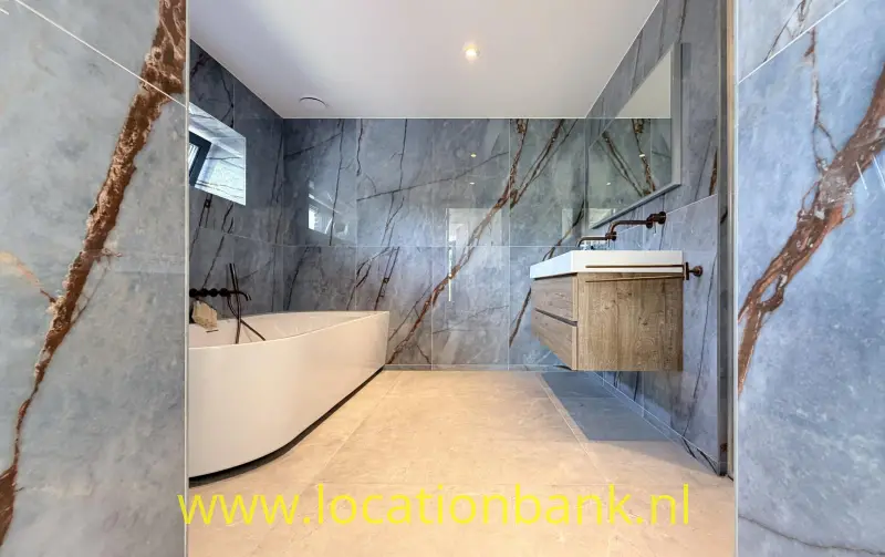 Badkamer met een vrijstaand wit bad, een houten wastafelmeubel en grijze wandtegels met bruine aders.
