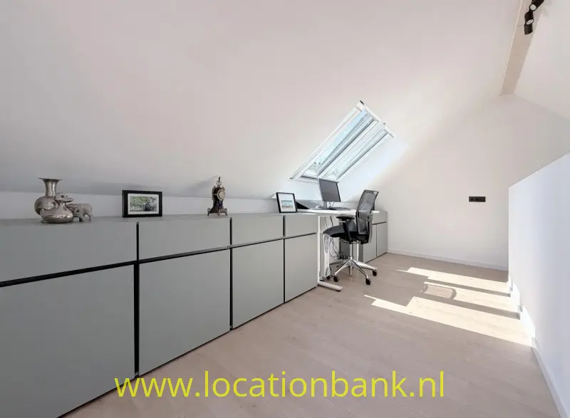 Lange lage kastenwand in een lichte zolderruimte met een dakraam, bureau en bureaustoel.