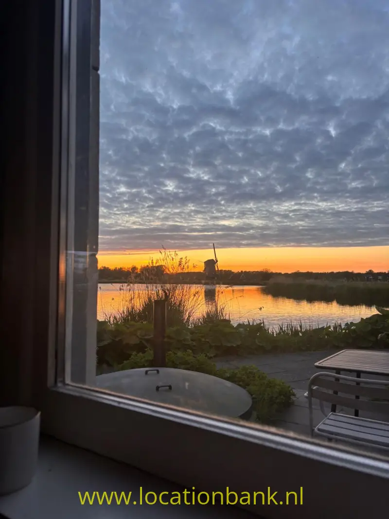 Een met riet omzoomd meer weerspiegelt de oranje zonsondergang, gezien door een raam.