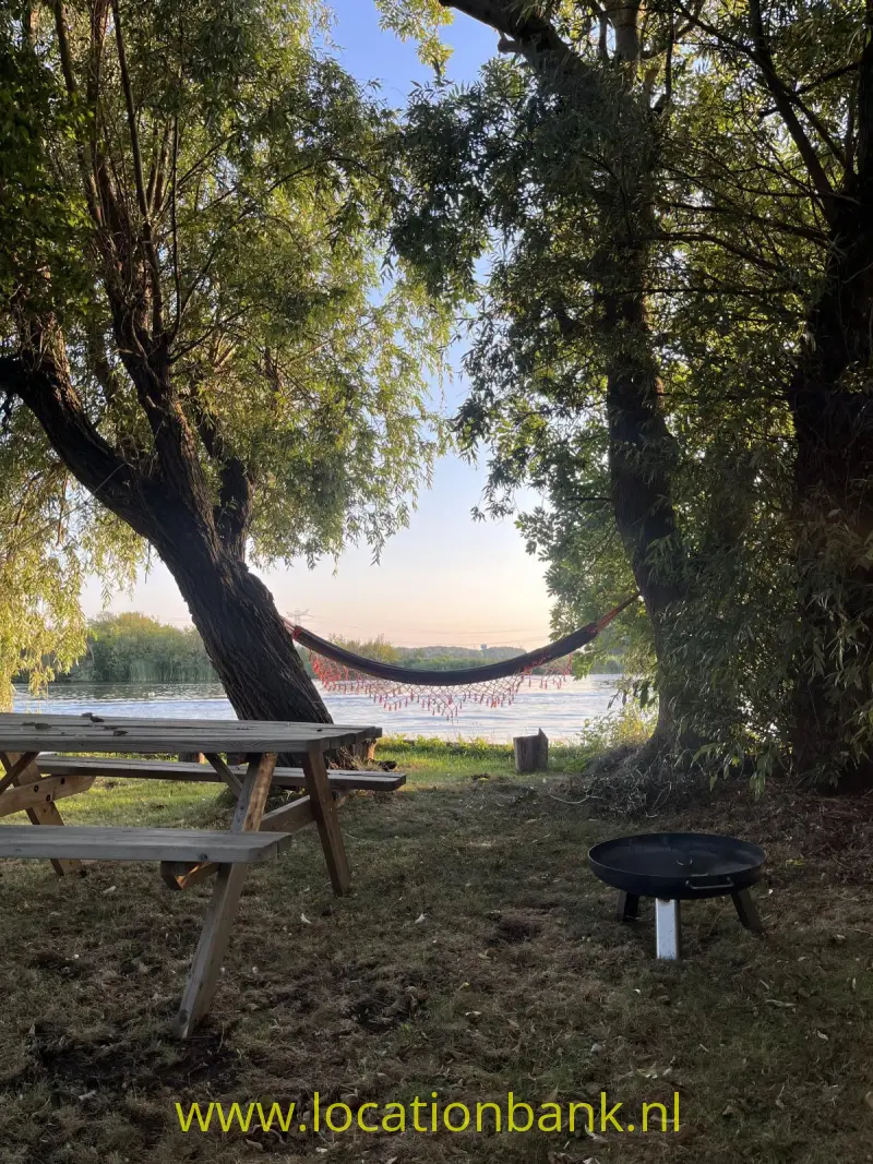 Een houten picknicktafel staat onder bomen in de tuin naast een hangmat aan de waterkant.
