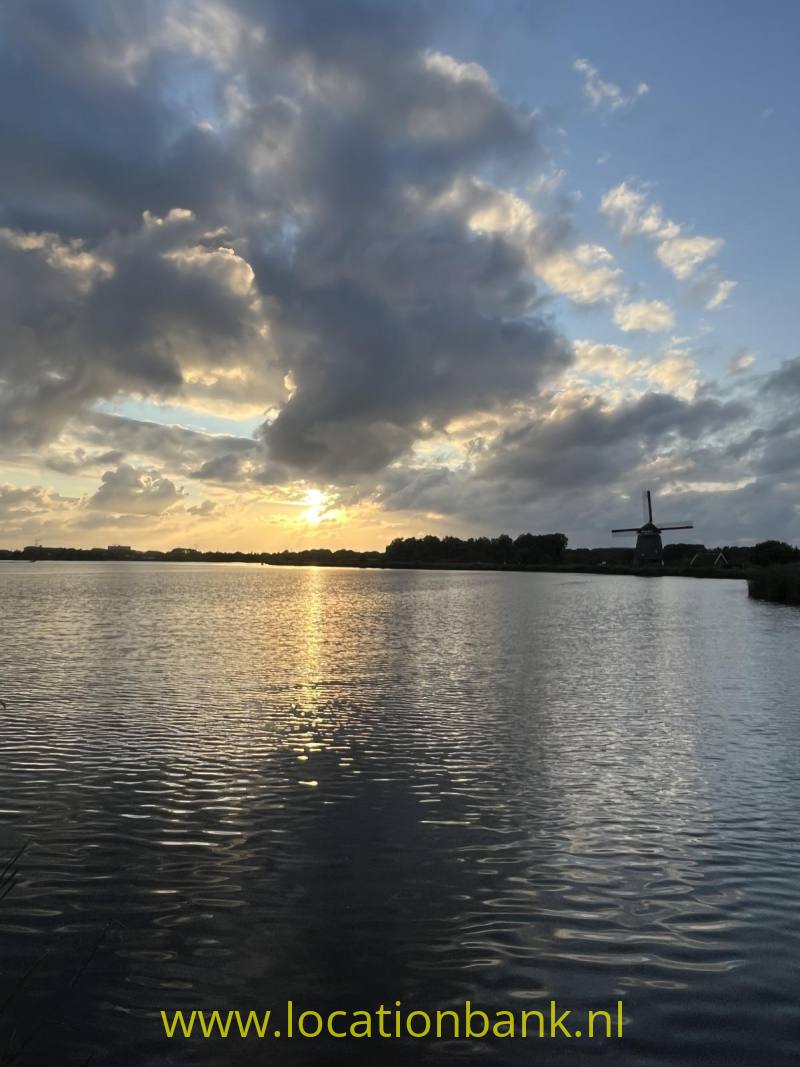 Een molen staat aan de waterkant, terwijl de laagstaande zon tussen donkere wolken door schijnt.