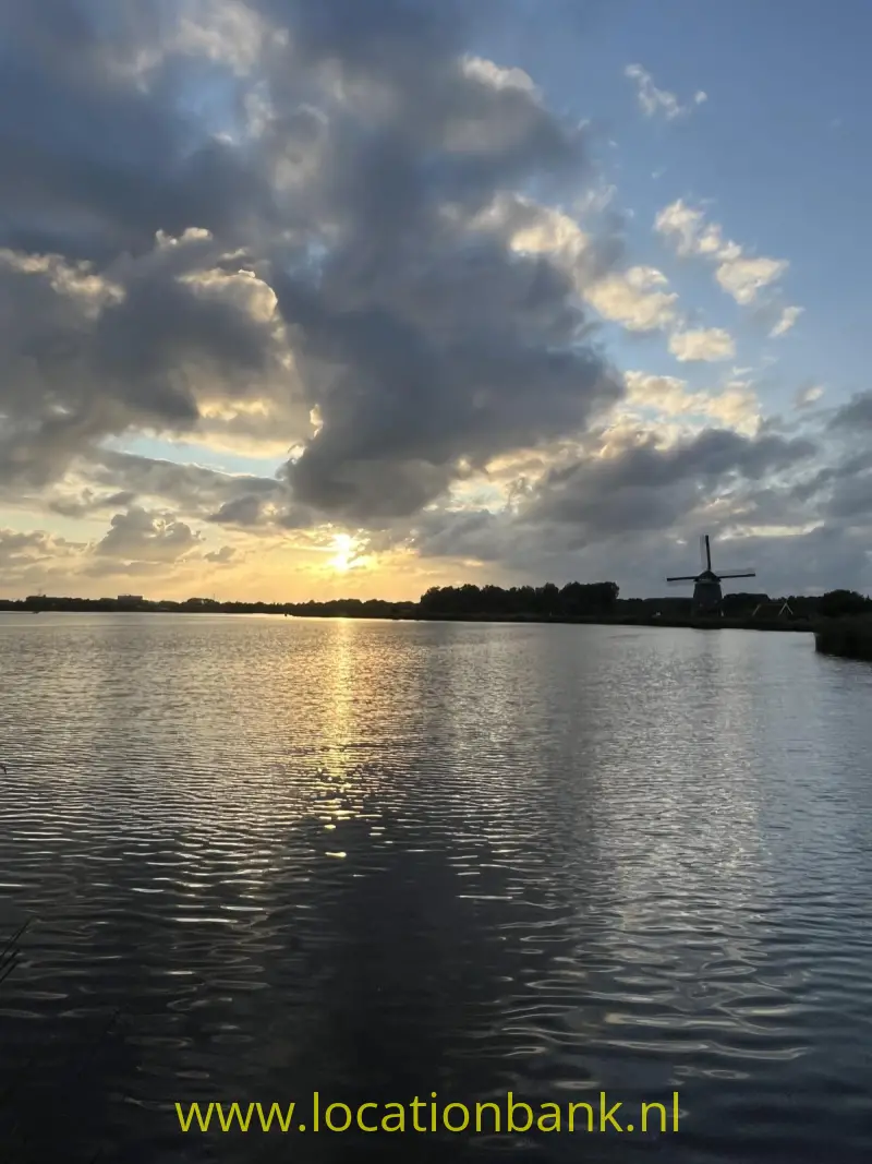 Een molen staat aan de waterkant, terwijl de laagstaande zon tussen donkere wolken door schijnt.