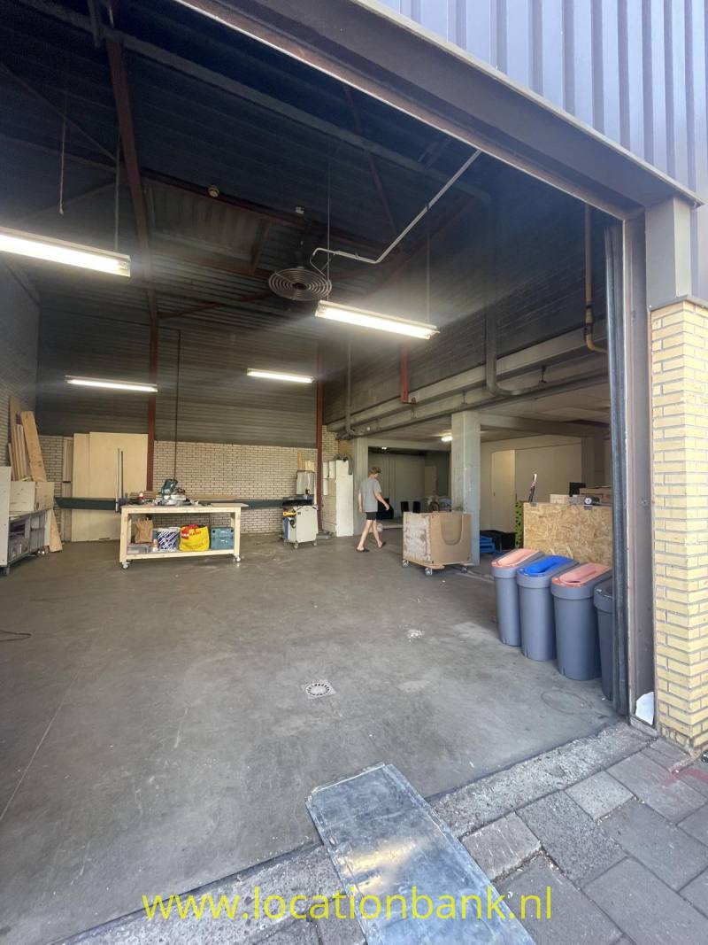 Een open hal met een metalen dakconstructie, werkbanken, afvalcontainers en een persoon bij de ingang.